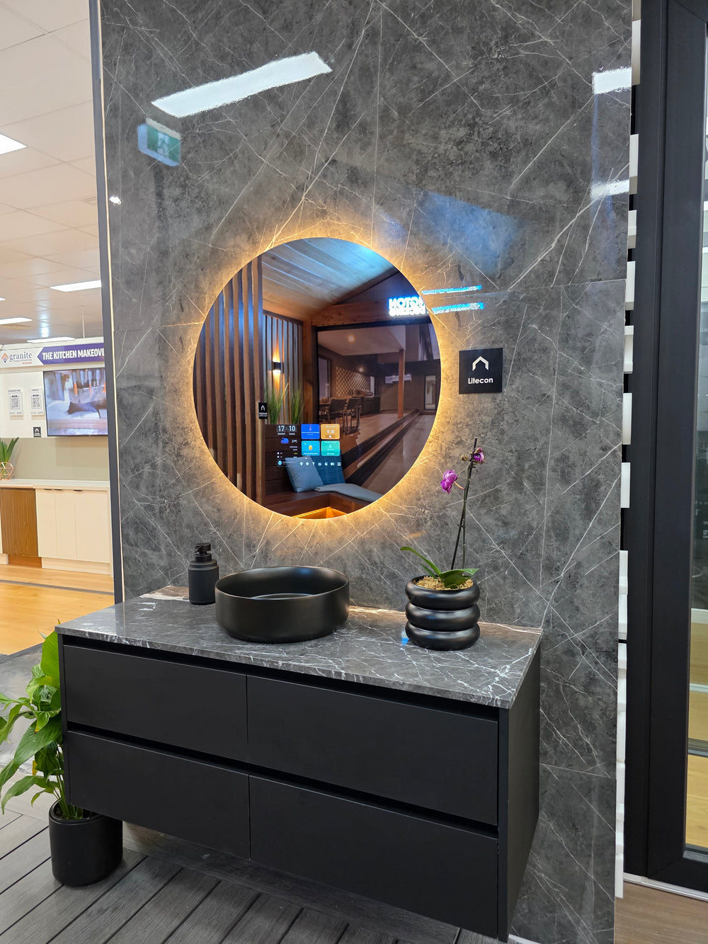 A modern floating bathroom vanity with a black drawer unit, a Gray marble-look counter top, a black vessel sink, and a large round back lit Litecon Smart mirror mounted on a dark Gray marble wall. The scene is photographed in a showroom with a reception desk visible in the background.