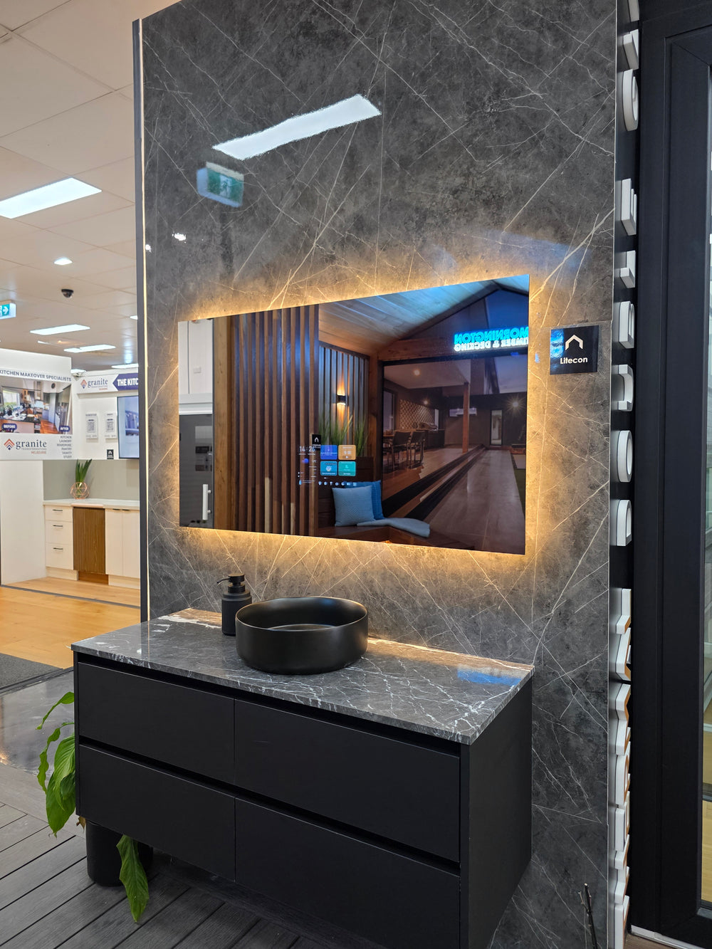 A modern bathroom vanity setup with a black floating cabinet, round black vessel sink, and a large backlit rectangular mirror mounted on a dark gray marble-look wall. The scene appears to be shown in a showroom or interior space, with floor, ceiling lights, and adjacent architectural elements visible.